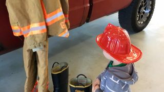 toddler wearing plastic fireman hat standing in front of fire truck looking at gear at touch-a-truck fire station open house